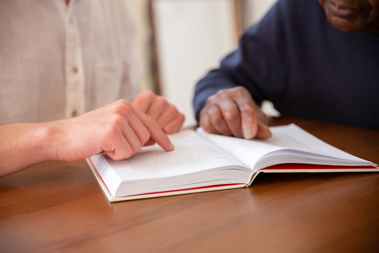 Close-up of two individuals reading a book together at a wooden table.