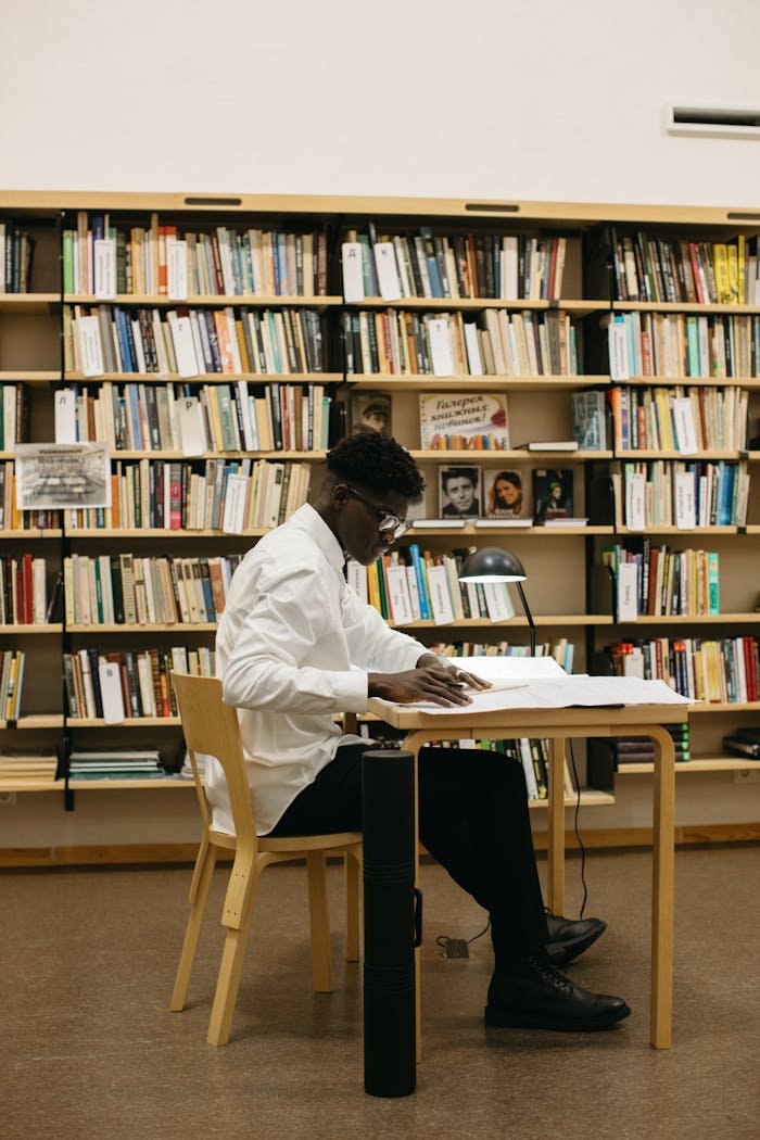 A determined student studying at a desk in a library full of bookshelves.