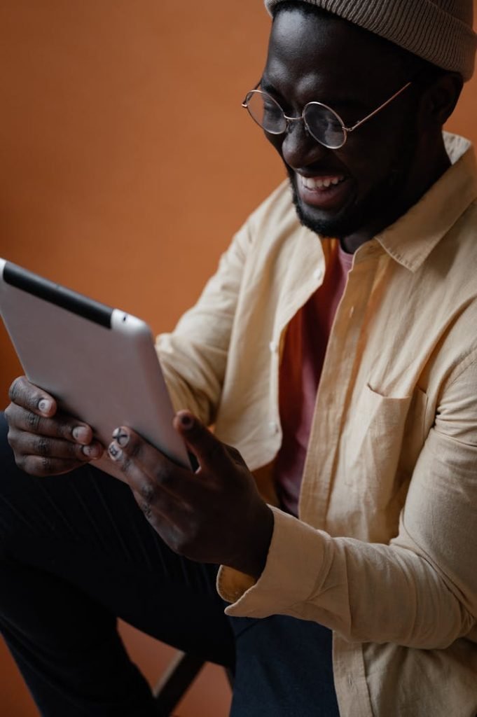 Cheerful African American male in casual clothes and glasses sitting on chair and using tablet against orange background