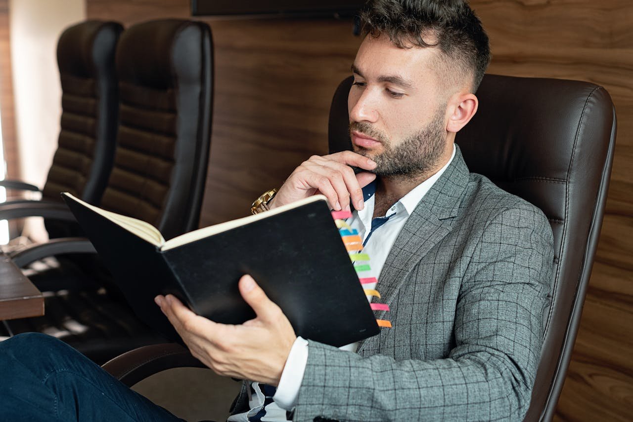 Businessman in suit reading notebook in modern office setting, planning and strategizing.