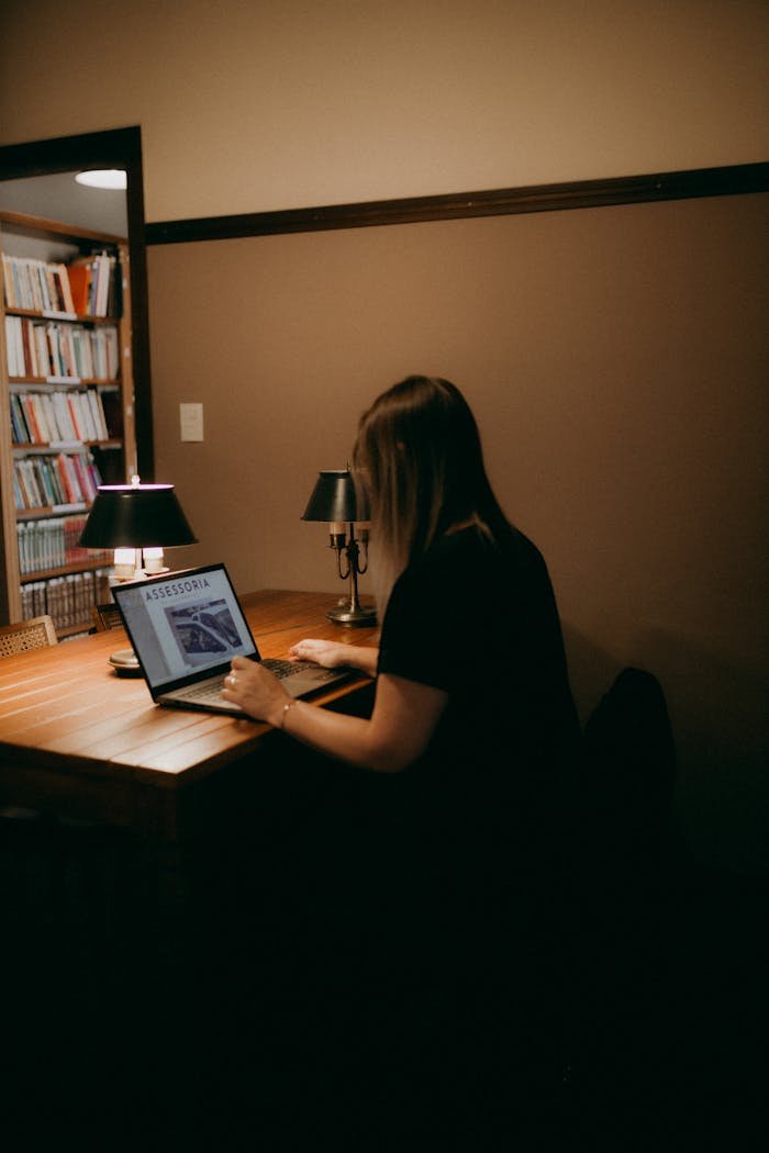 A woman works on her laptop in a warm, cozy home office with bookshelves and soft lighting.